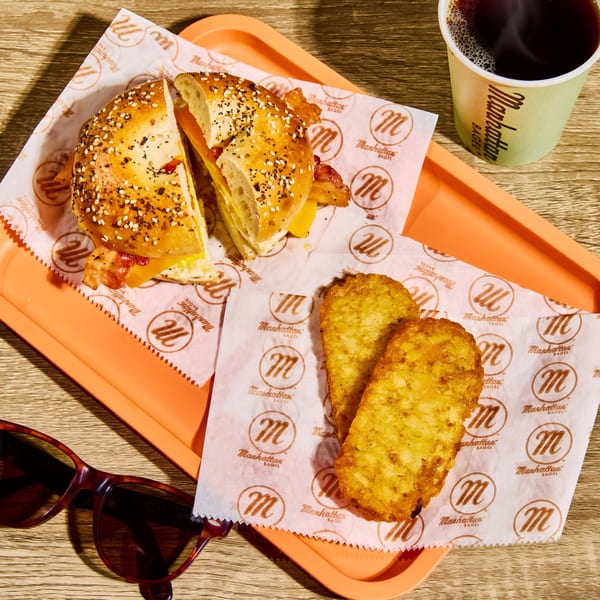 Manhattan Bagel breakfast sandwich, hash browns, and coffee on an orange tray with sunglasses.