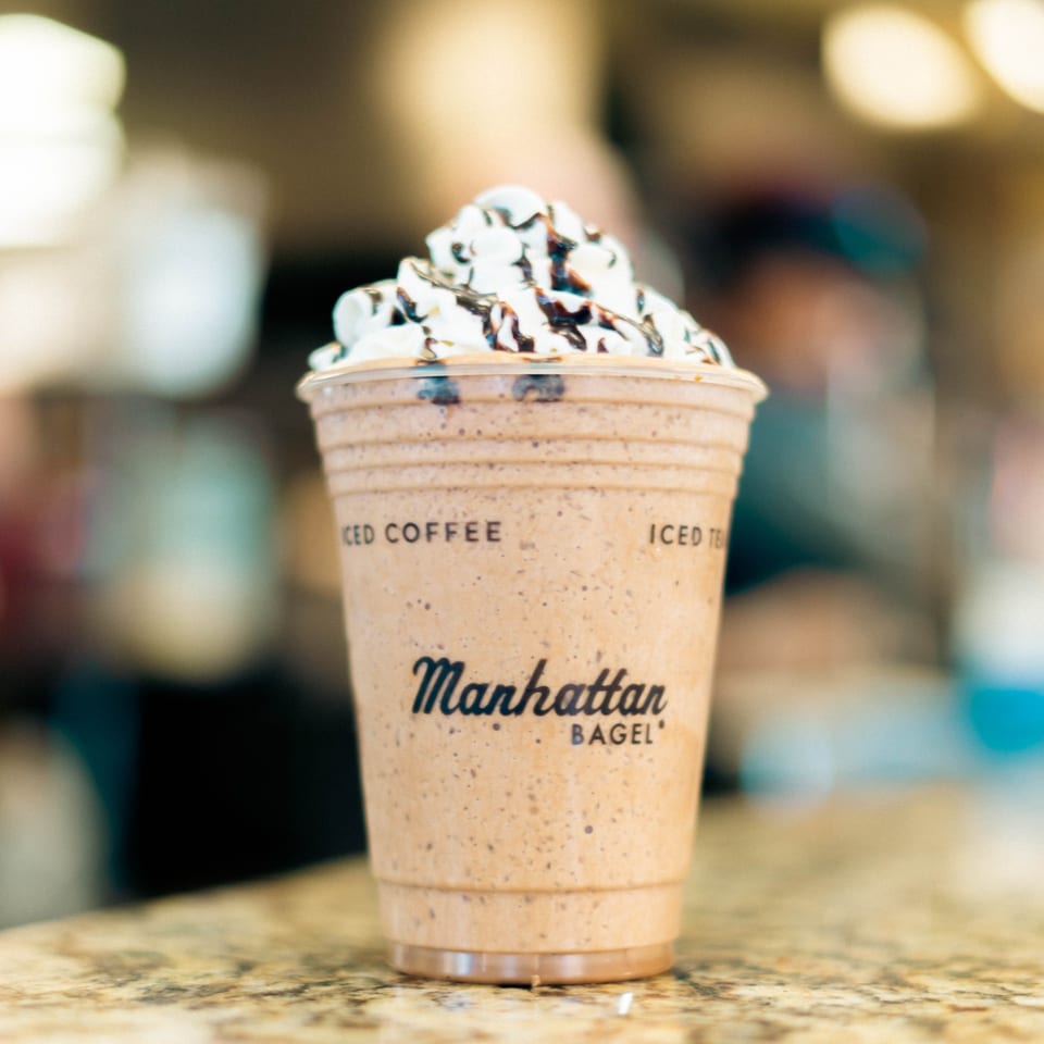 A Manhattan Bagel iced coffee drink with whipped cream and chocolate drizzle on a counter.