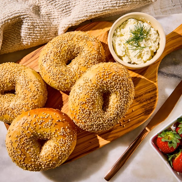 Four sesame bagels, a bowl of cream cheese with dill, and fresh strawberries on a wooden board.
